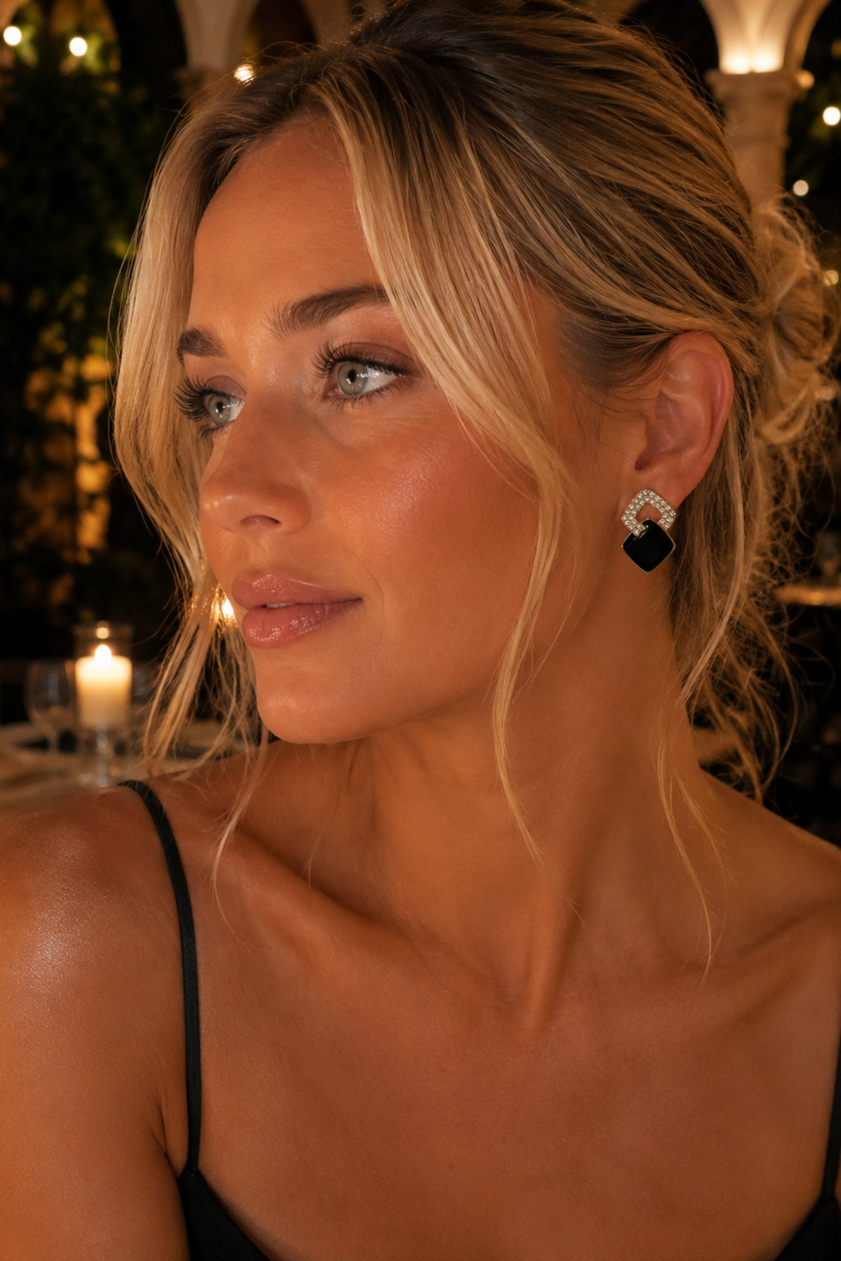 A close-up portrait of a woman with blonde hair in an updo, wearing a black spaghetti-strap top and a black and silver geometric stud earring, set against a warm, softly blurred background with candles.
