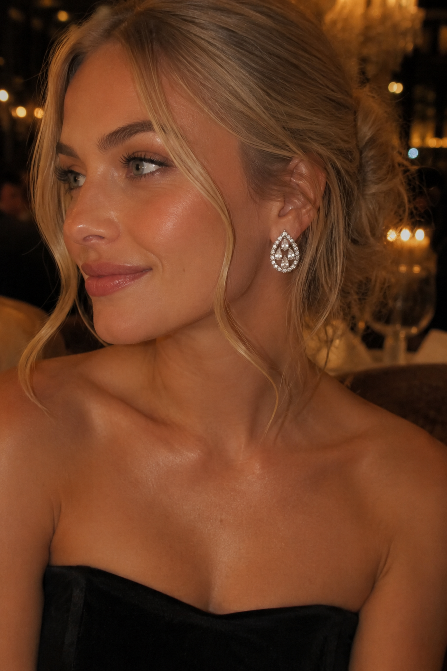 A close-up portrait of a woman with blonde hair in an updo, wearing a sparkling teardrop-shaped diamond drop earring and a black strapless top, set against a warm, softly blurred background with candlelight.