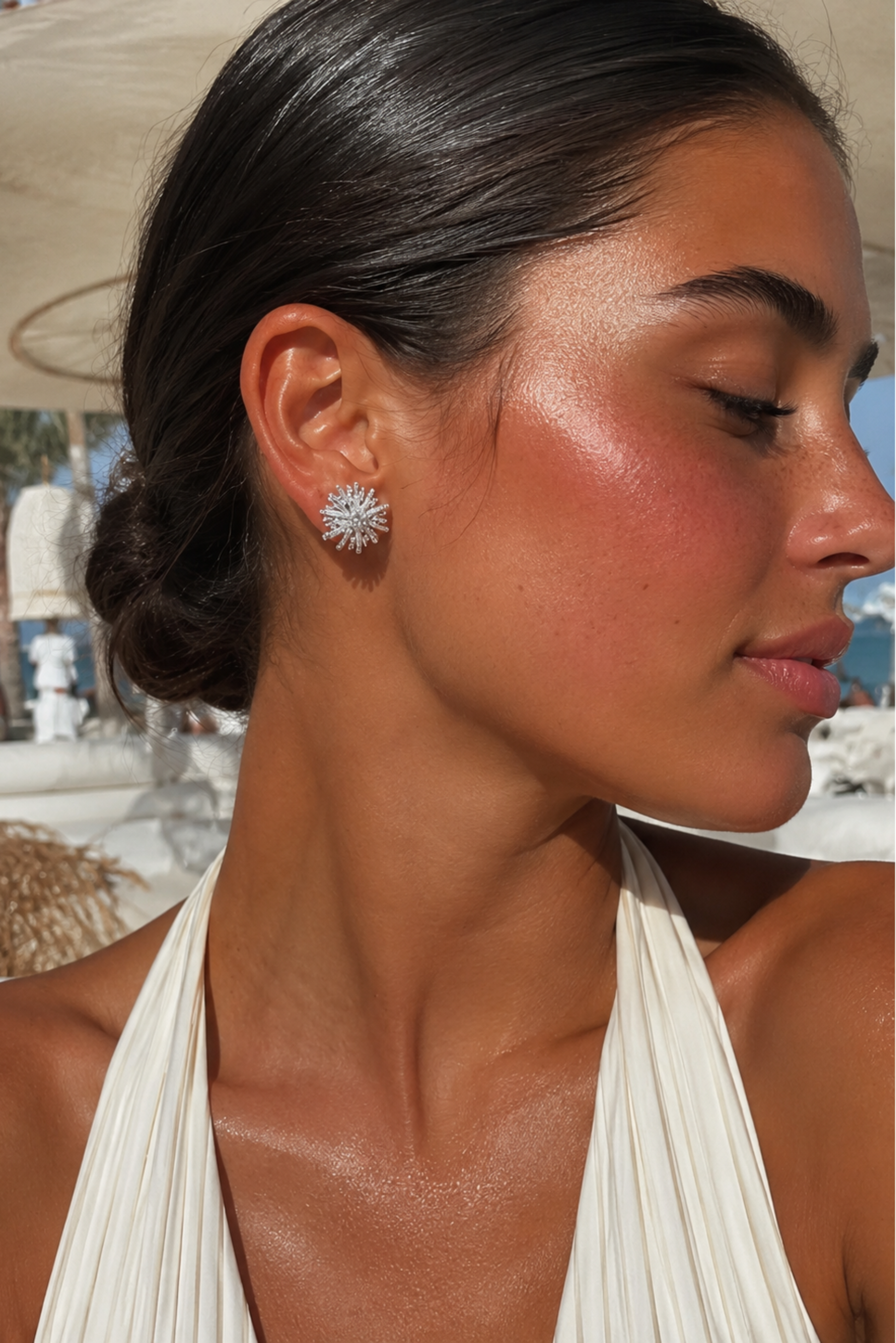 A close-up side profile of a woman with dark hair in a low bun, wearing a silver-toned starburst stud earring and a cream halter top, set against a sunny beach resort background.