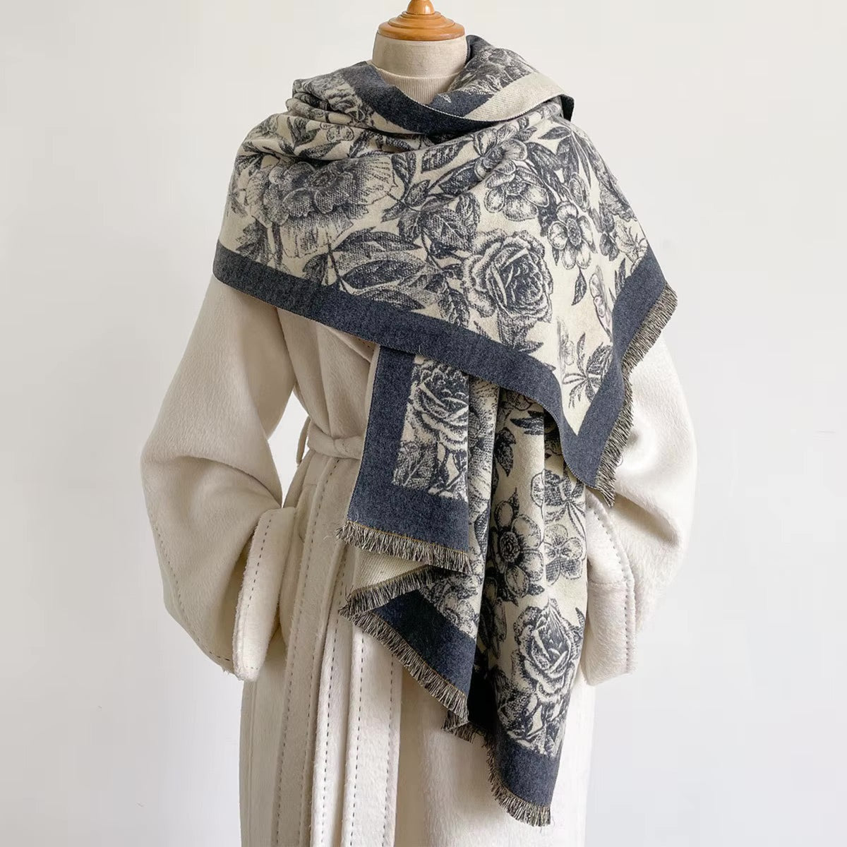A cream-colored shawl with a dark blue floral toile pattern and fringed edges, draped over a cream-colored coat on a mannequin torso against a plain white background.