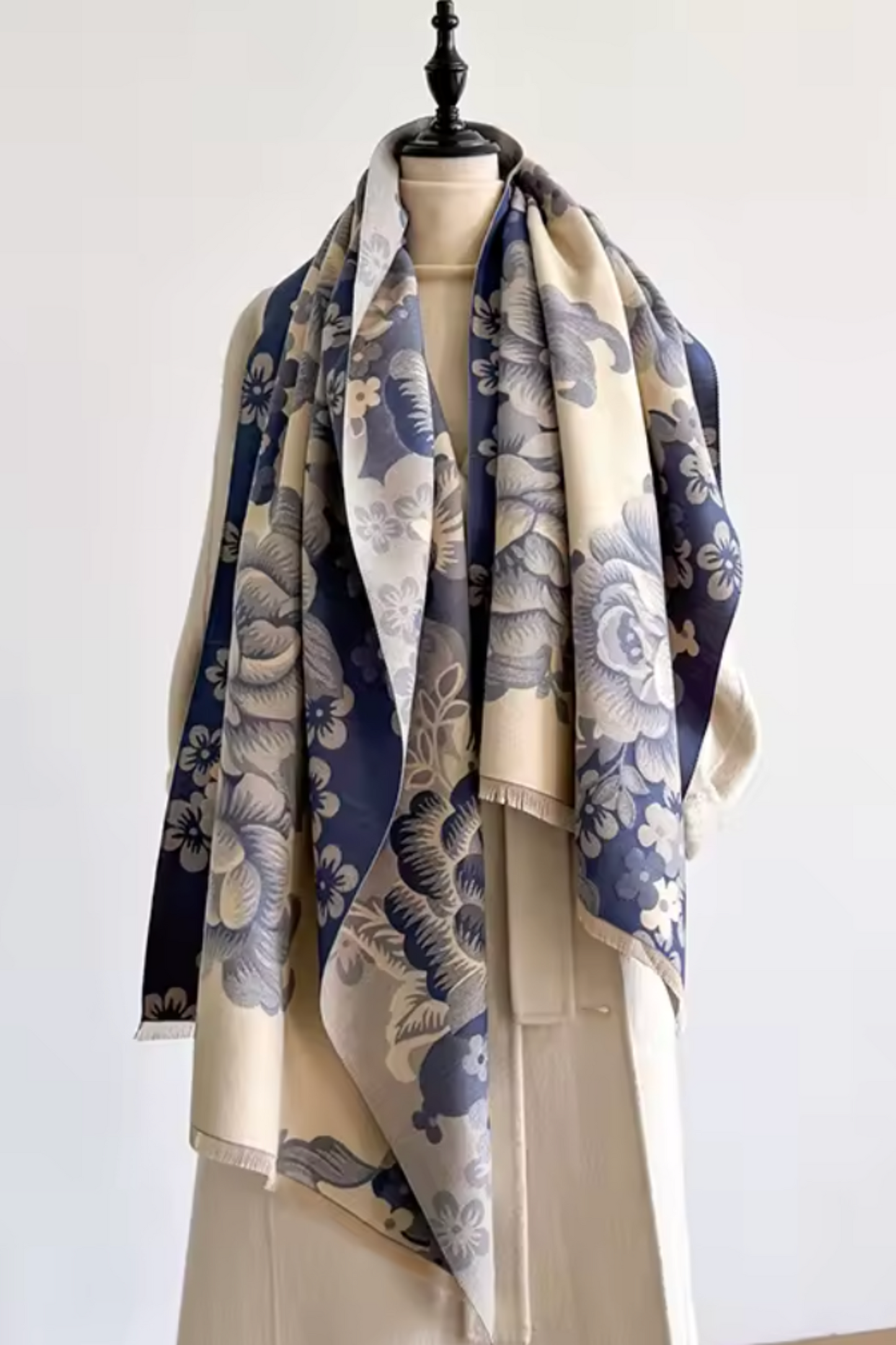 A cream-colored silk scarf with a navy blue and gray floral print, elegantly draped on a cream-colored mannequin dress form against a plain white wall.