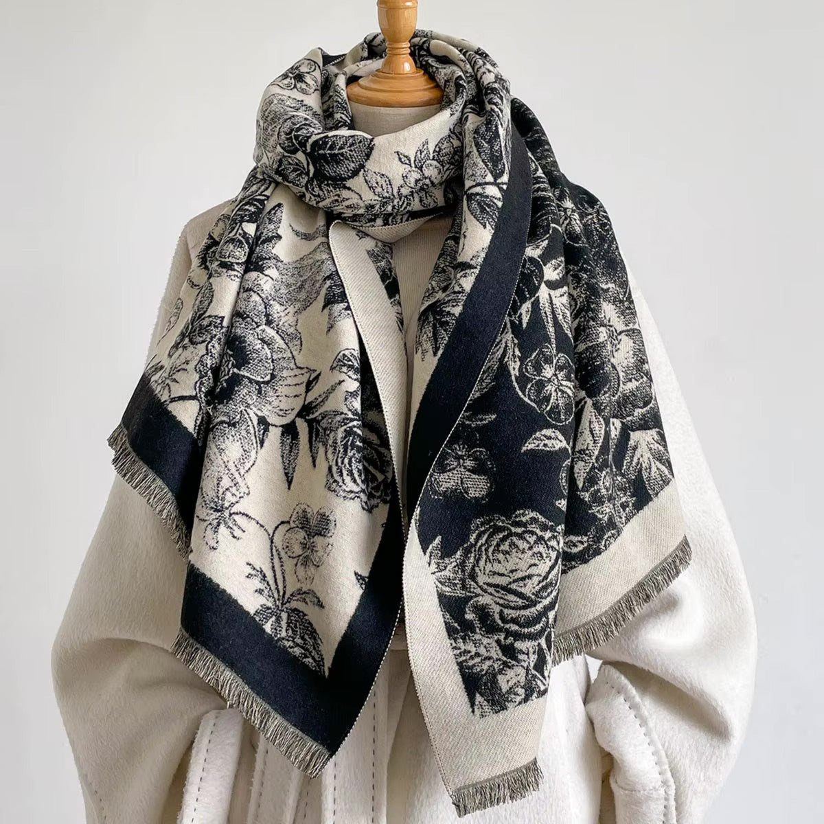 A cream and navy blue patterned scarf with fringed edges, draped on a mannequin wearing a cream coat against a plain light background.