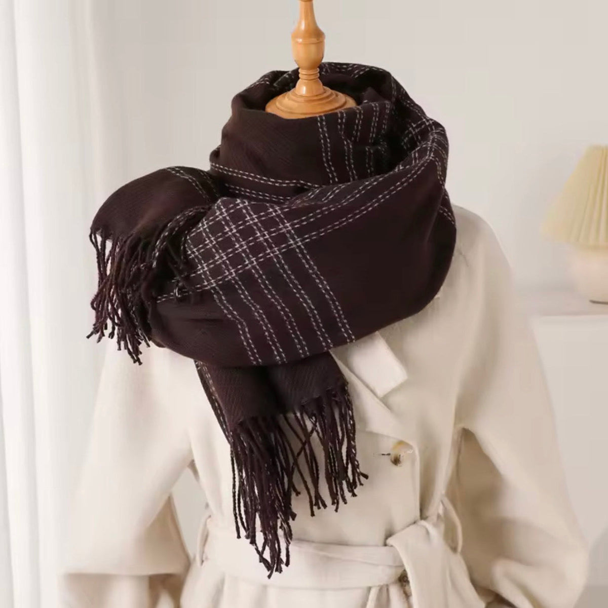 A dark brown plaid scarf with white stitching and fringed ends, wrapped around a cream-colored coat displayed on a wooden mannequin torso.