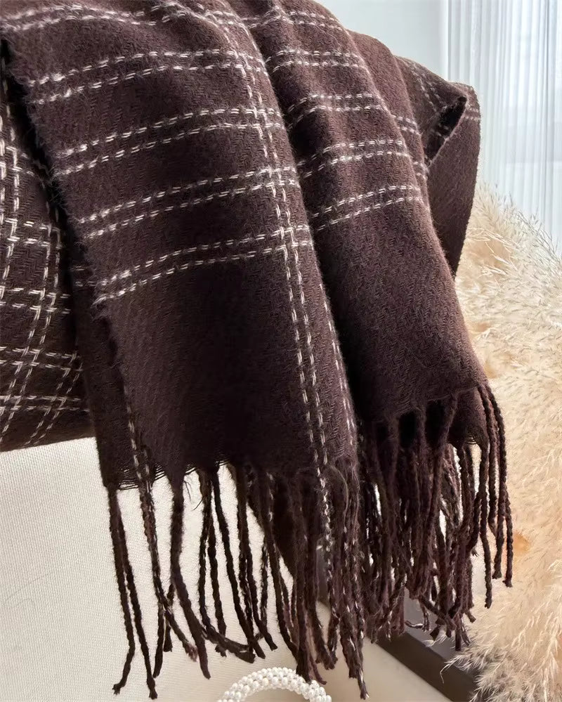 A dark brown plaid throw blanket with white grid lines and long fringe tassels, draped over a light-colored surface next to a fluffy beige faux-fur accessory.