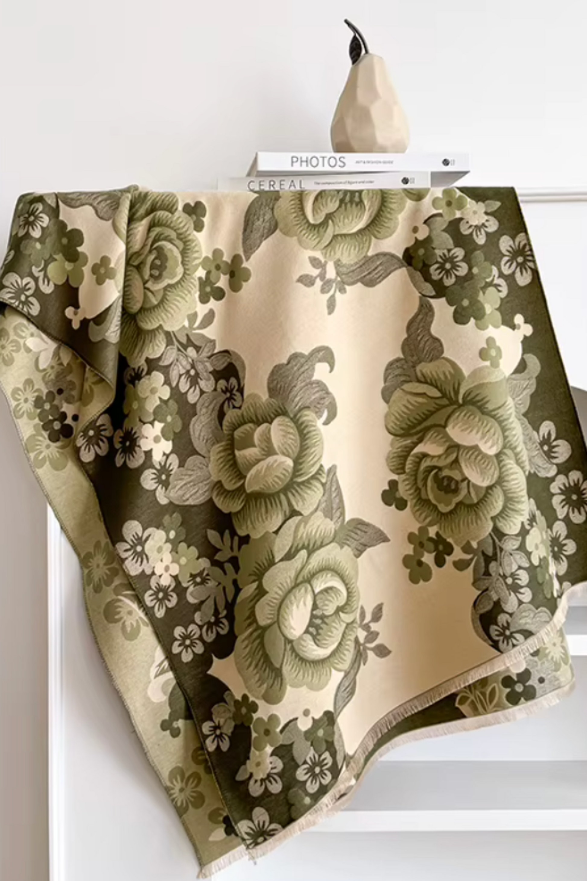 A draped fabric throw or scarf with a large floral print in olive green and cream, styled over a white shelf with books and a decorative object.