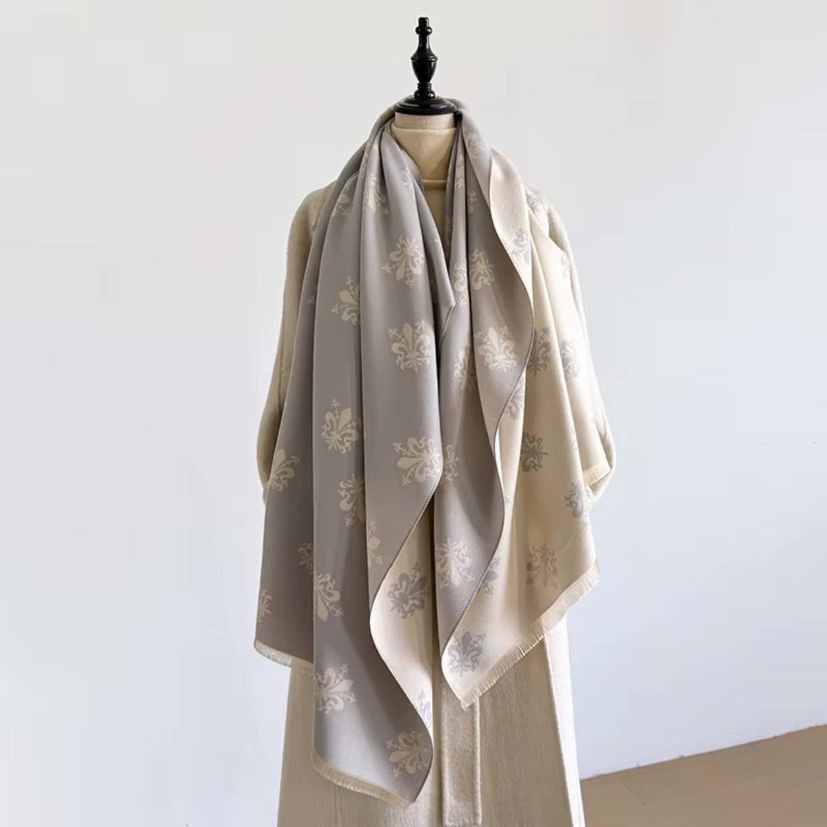 A draped scarf in warm beige and light gray with woven floral motifs, displayed on a mannequin against a plain white wall.