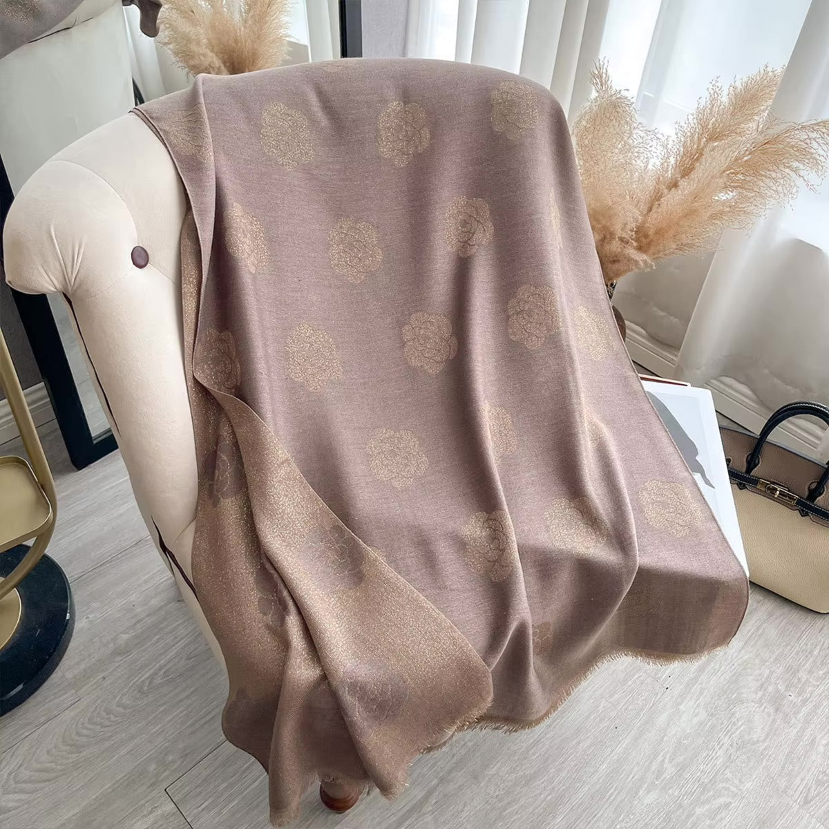 A draped throw blanket in a dusty taupe-brown color with a repeating tonal floral pattern, styled over a cream tufted armchair in a bright room.
