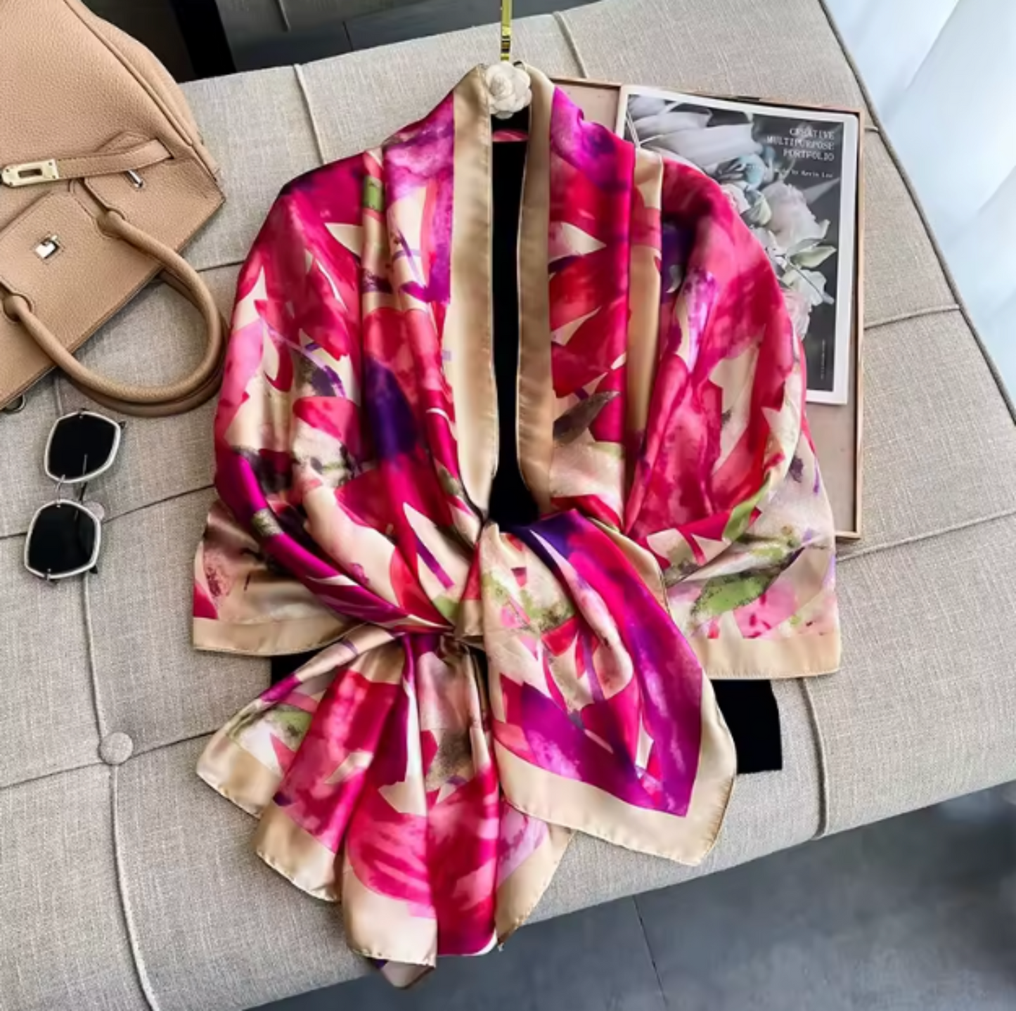 A floral silk scarf with a vibrant pink, magenta, and purple watercolor-style print on a beige base, styled on a gray upholstered bench with a handbag, sunglasses, and a book.