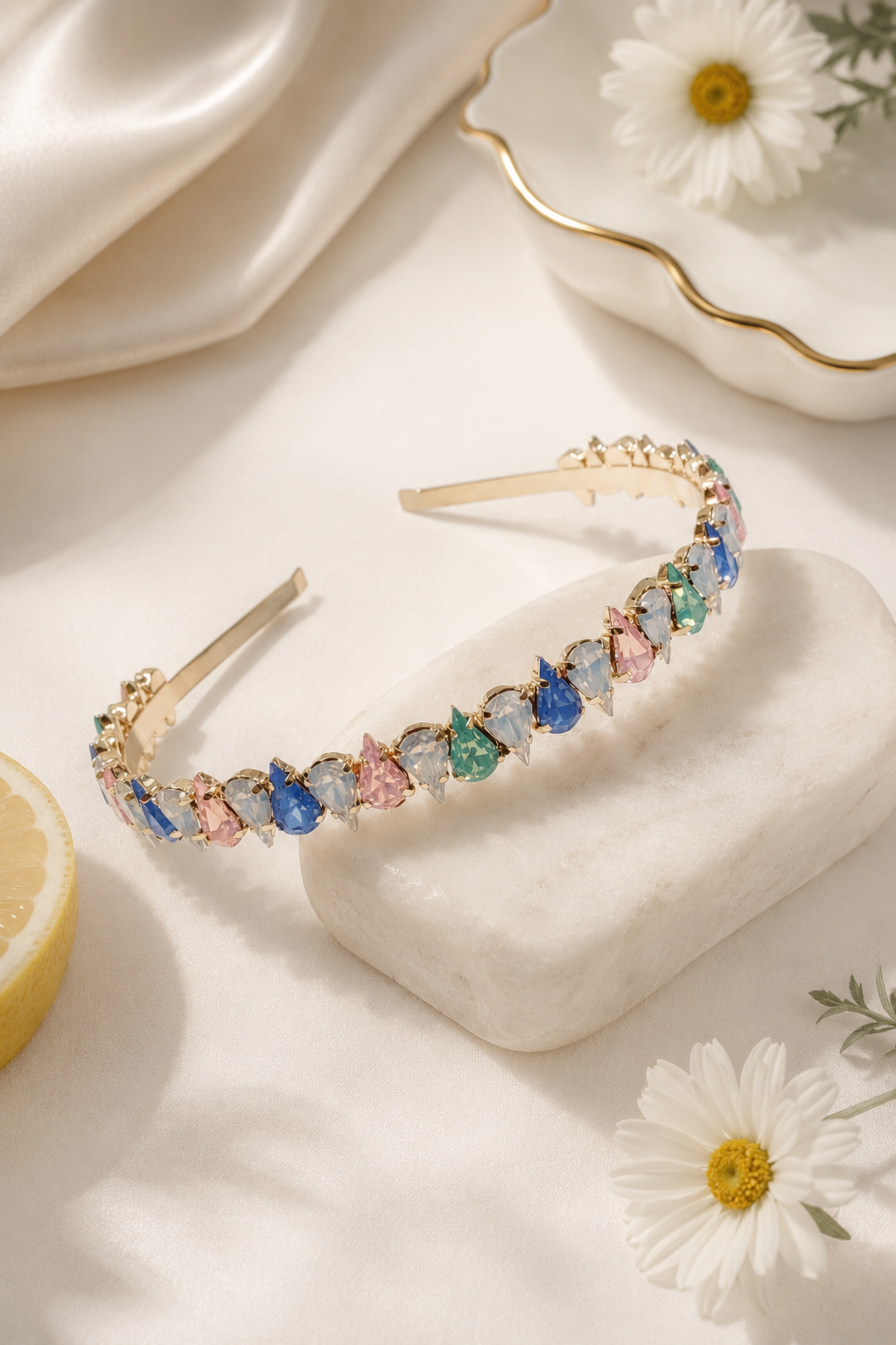 A gold-tone headband adorned with colorful teardrop-shaped rhinestones in shades of blue, green, pink, and white, styled on a white marble block with daisies and a lemon slice.