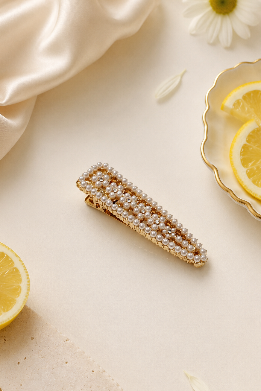 A gold-toned hair claw clip embellished with rows of white faux pearls, styled in a flat-lay composition with lemon slices, white daisies, a gold chain, and cream-colored satin fabric.