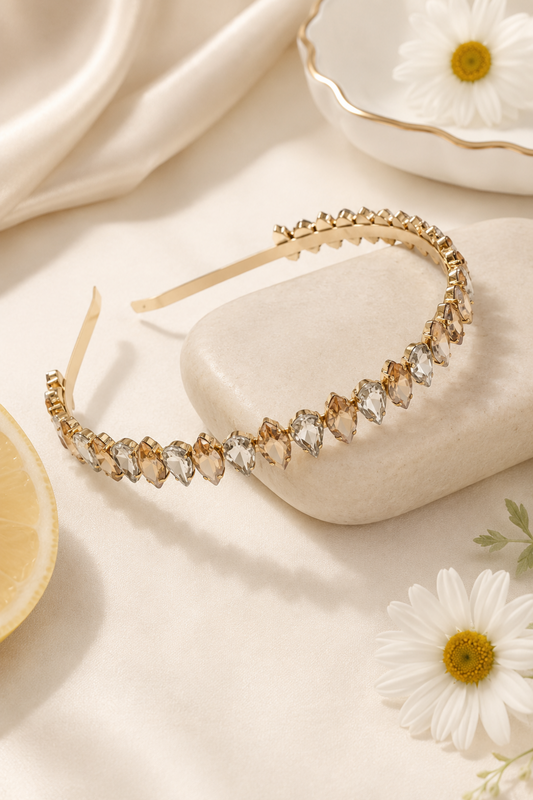 A gold headband adorned with clear teardrop-shaped crystals, styled on a cream-colored stone with a lemon slice, white daisies, and draped satin fabric in the background.