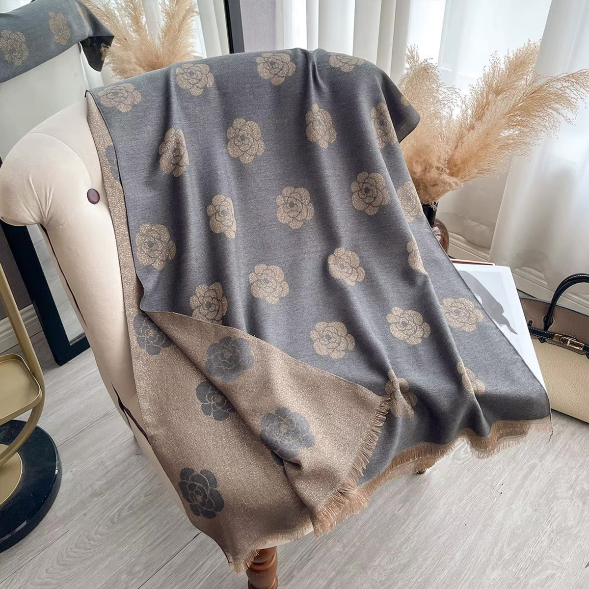A gray and beige patterned throw blanket with a floral camellia design is draped over a cream-colored armchair in a bright, modern room setting.