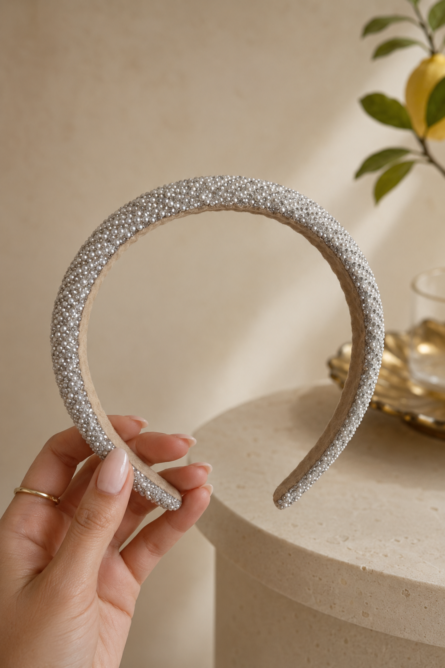 A hand holds a wide headband covered in sparkling silver rhinestones, set against a warm beige background with a decorative pedestal and lemon branch.