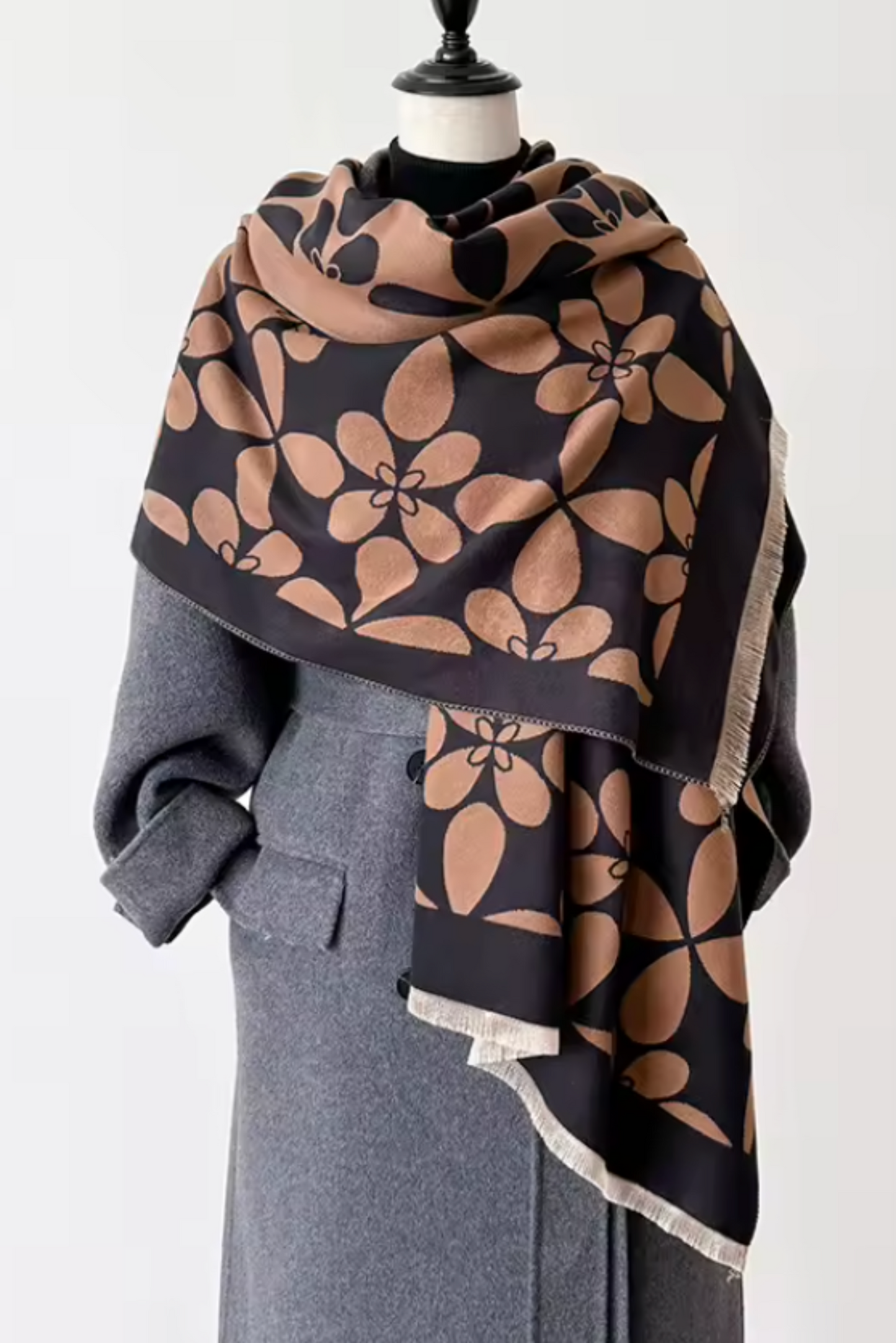 A large patterned scarf draped over a gray coat on a mannequin torso against a plain light background. The scarf has a dark charcoal base with large tan floral motifs and a light beige fringe edge.