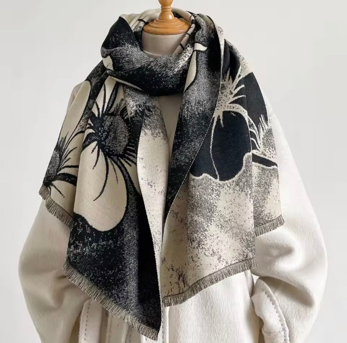A large square scarf with a black and beige floral print, draped on a cream-colored coat displayed on a mannequin torso against a plain light background.