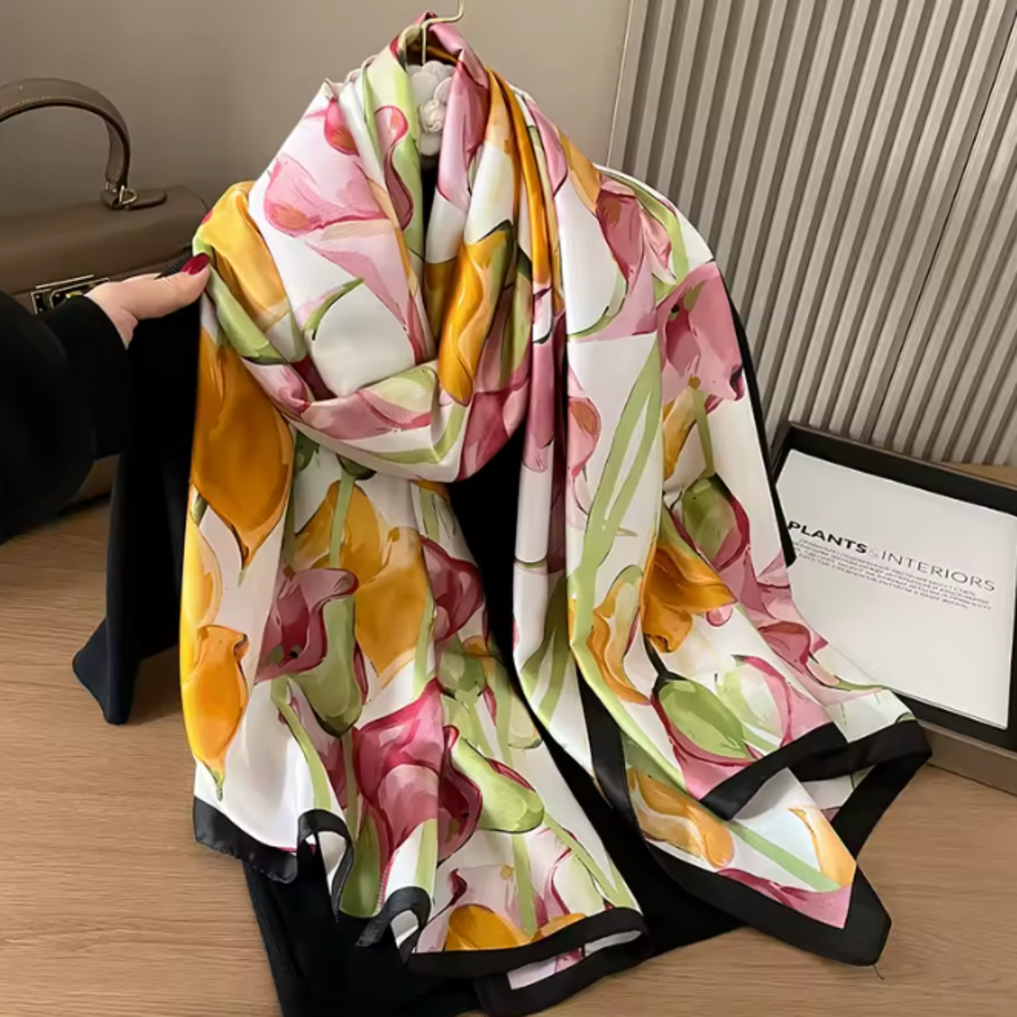 A large square scarf with a colorful abstract floral print in pink, yellow, green, and orange on a white background, styled with black garments and a framed print on a wooden floor.