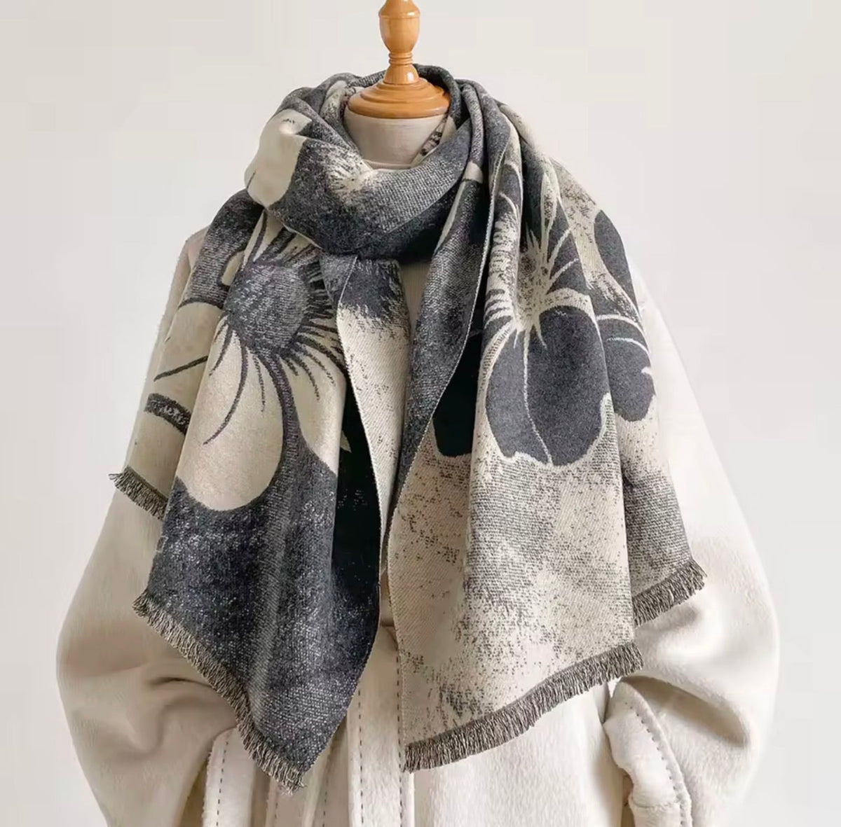 A large square scarf with a distressed floral pattern in charcoal gray and cream, styled on a wooden mannequin torso wearing a cream coat against a plain light background.