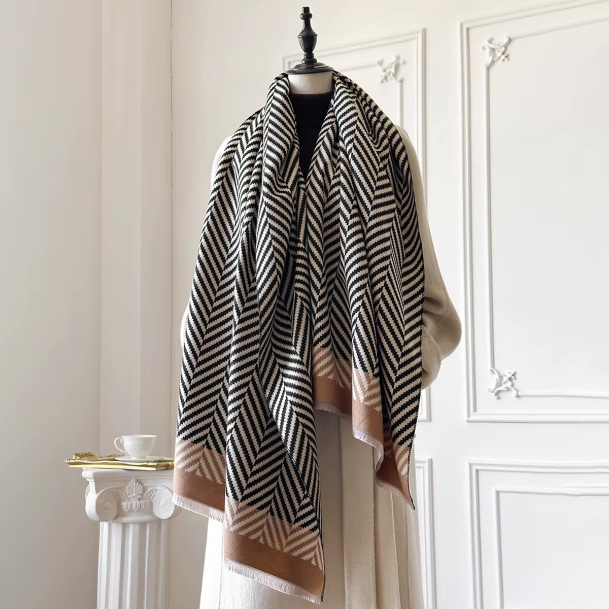 A large woven scarf with a black and beige herringbone pattern and tan borders, draped over a cream-colored coat on a mannequin in an elegant indoor setting.