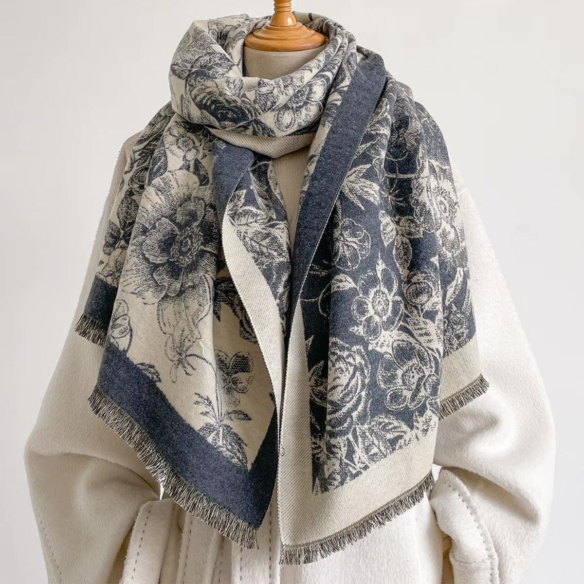 A large woven scarf with a floral toile pattern in navy blue and cream, draped on a cream-colored coat displayed on a mannequin torso against a plain white background.