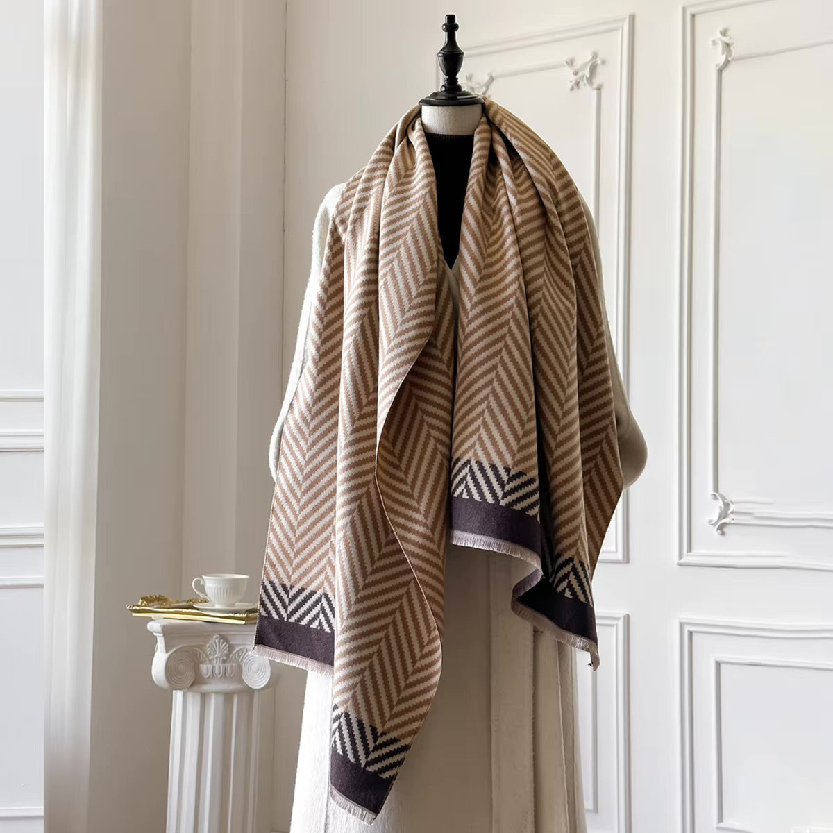 A large woven shawl with a beige and brown chevron pattern and dark purple borders, draped over a cream-colored dress form in an elegant room with white paneled walls.