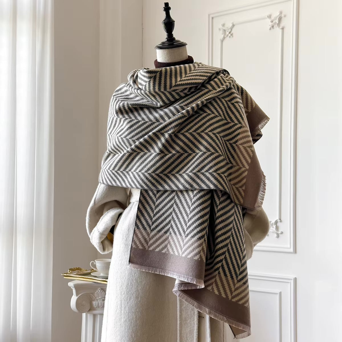 A large woven shawl with a beige and dark brown herringbone pattern draped over a cream-colored coat on a mannequin, set against a white paneled wall.