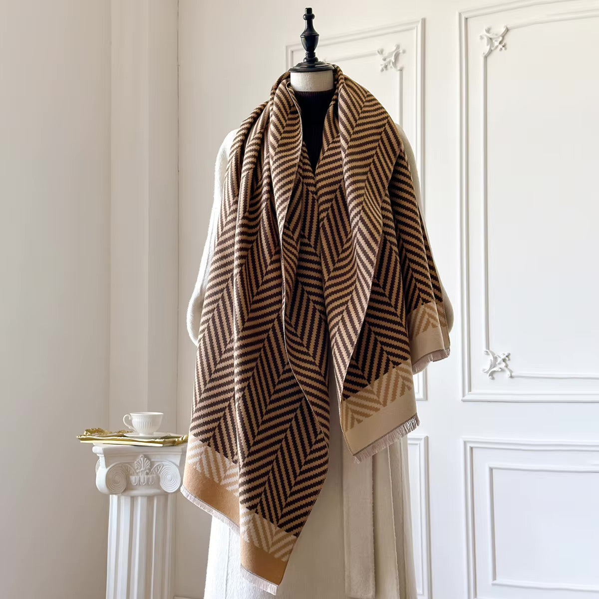 A large woven shawl with a brown and beige geometric chevron pattern and fringed edges, draped over a cream-colored dress form in an elegant room with white paneled walls.