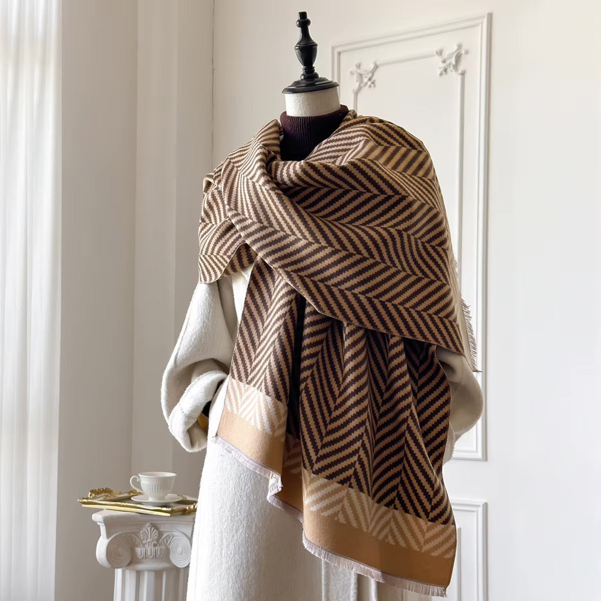 A large woven shawl with a brown and beige herringbone pattern and a solid tan border, draped over a cream-colored sweater on a mannequin in a bright, white room.