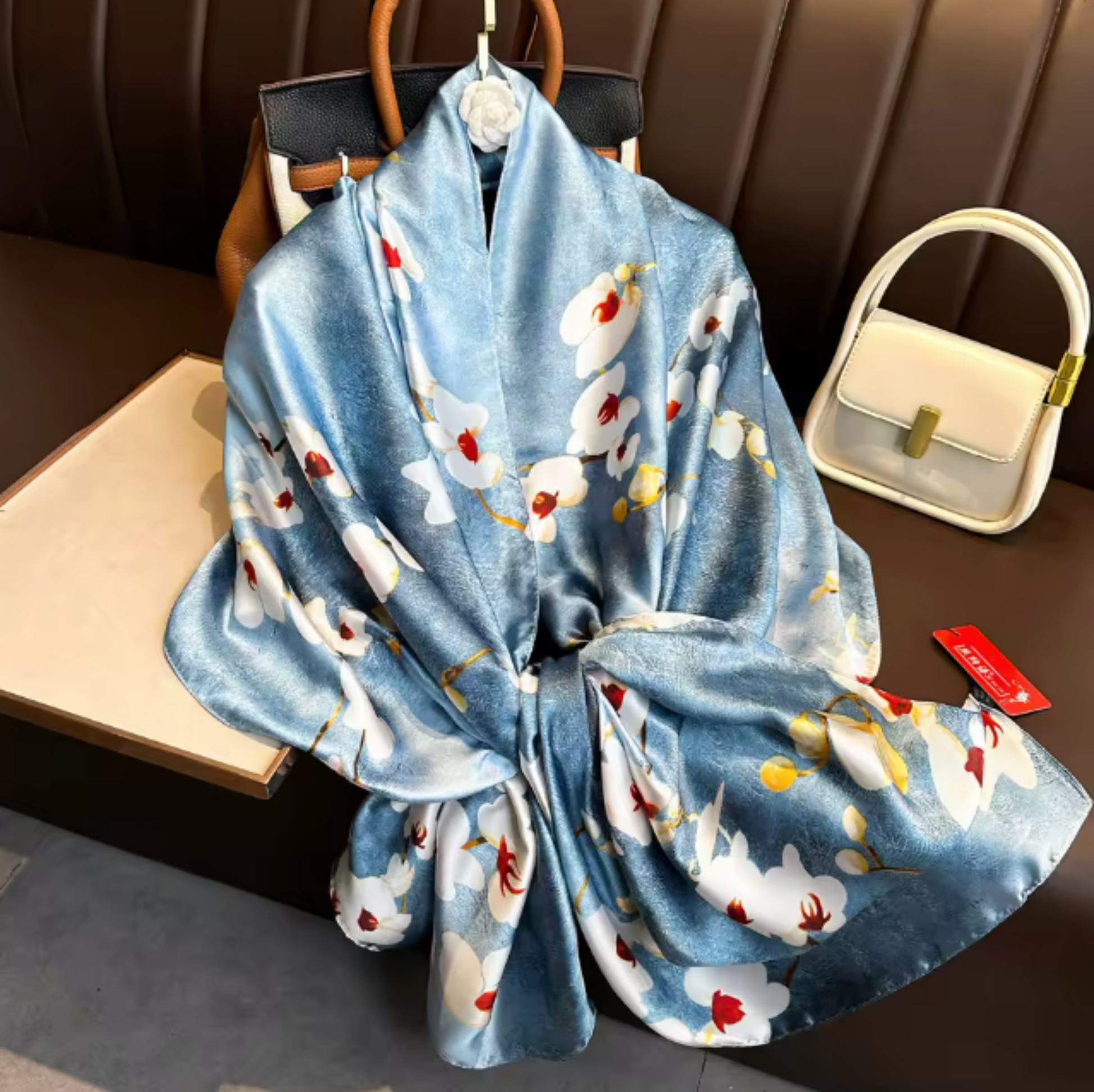 A light blue kimono-style robe with a white floral print and small red and yellow accents, draped over a hanger on a dark brown bench. A cream-colored handbag sits to the right, and a red tag is visible near the hem.