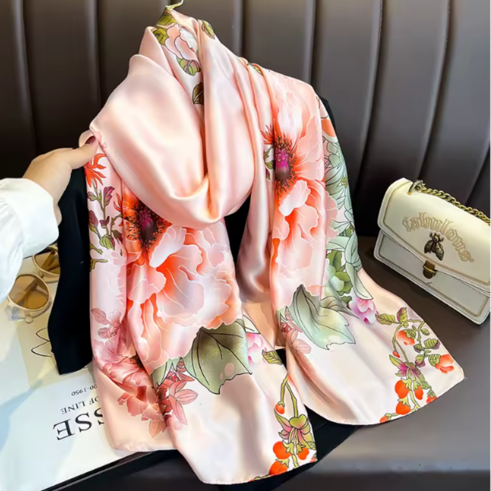 A light pink satin scarf with a large floral print in shades of orange, green, and purple, draped over a black leather chair next to a cream-colored handbag.