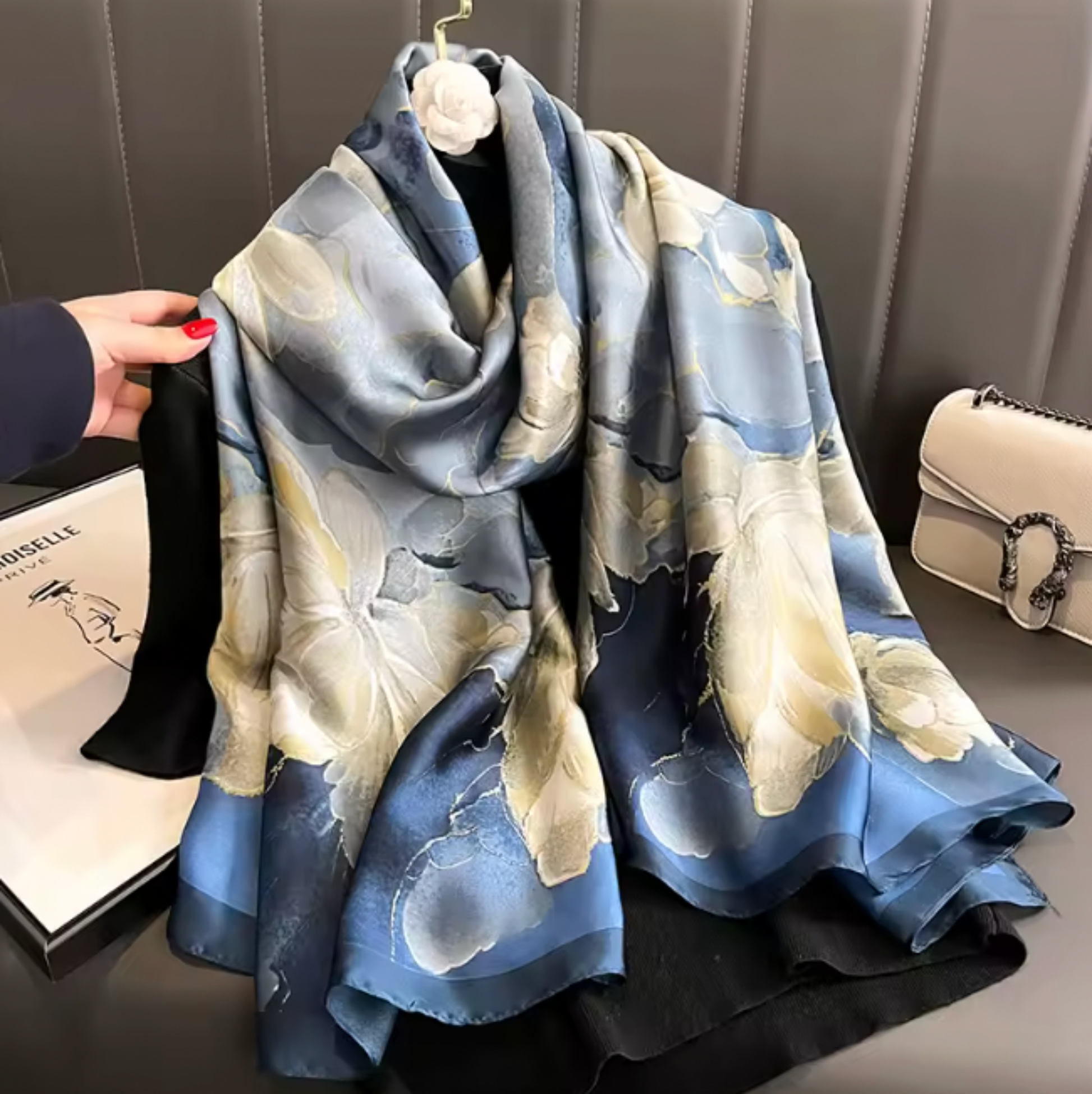 A lightweight square scarf with a blue and cream floral watercolor pattern, draped over a black garment on a hanger. A hand with red nail polish is holding the left edge of the scarf, next to a white Chanel box and a white Gucci shoulder bag on a dark gray leather surface.
