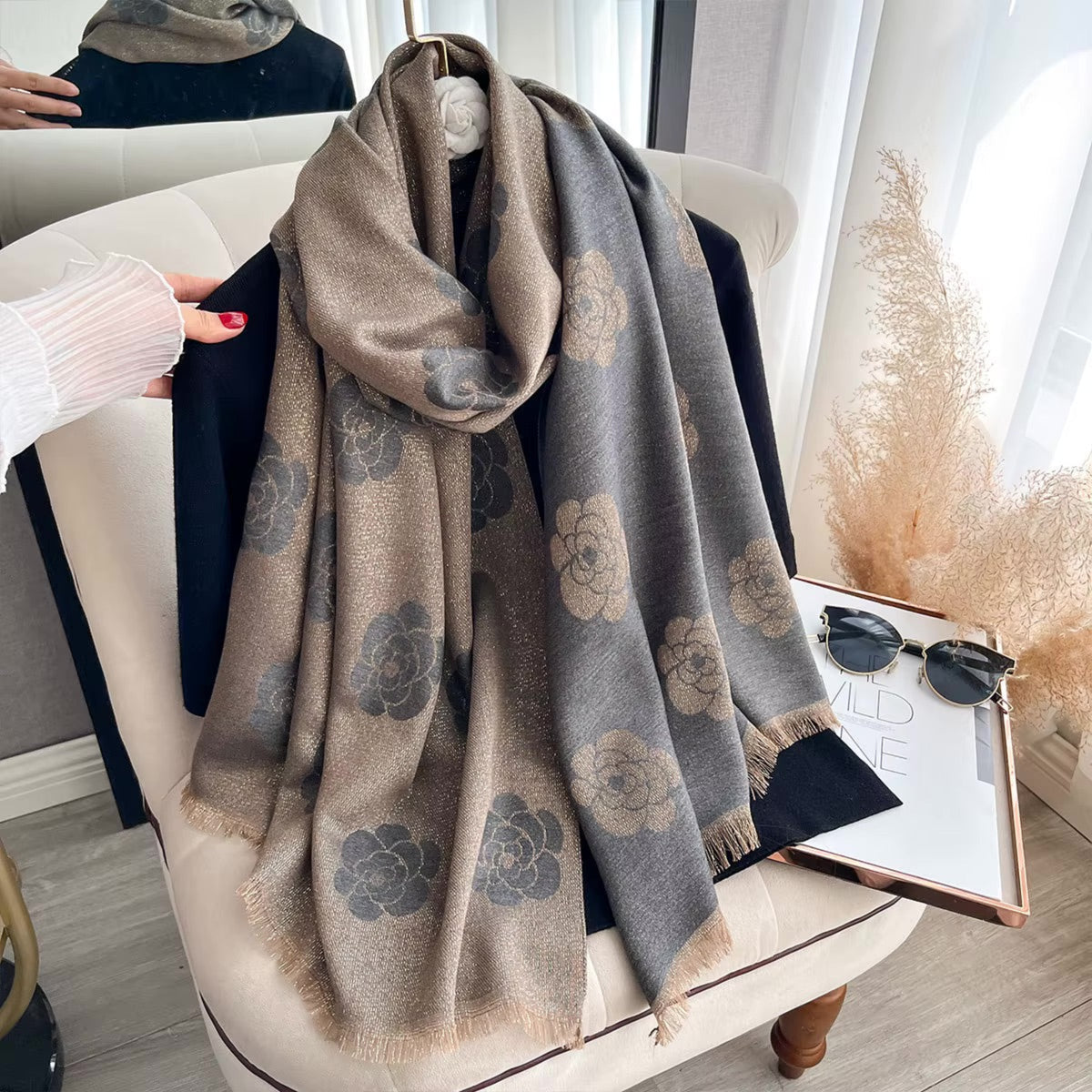 A long woven scarf with a large floral pattern, draped over a cream-colored armchair in a styled room. The scarf has a warm taupe-beige base with dark gray floral motifs and fringed edges.