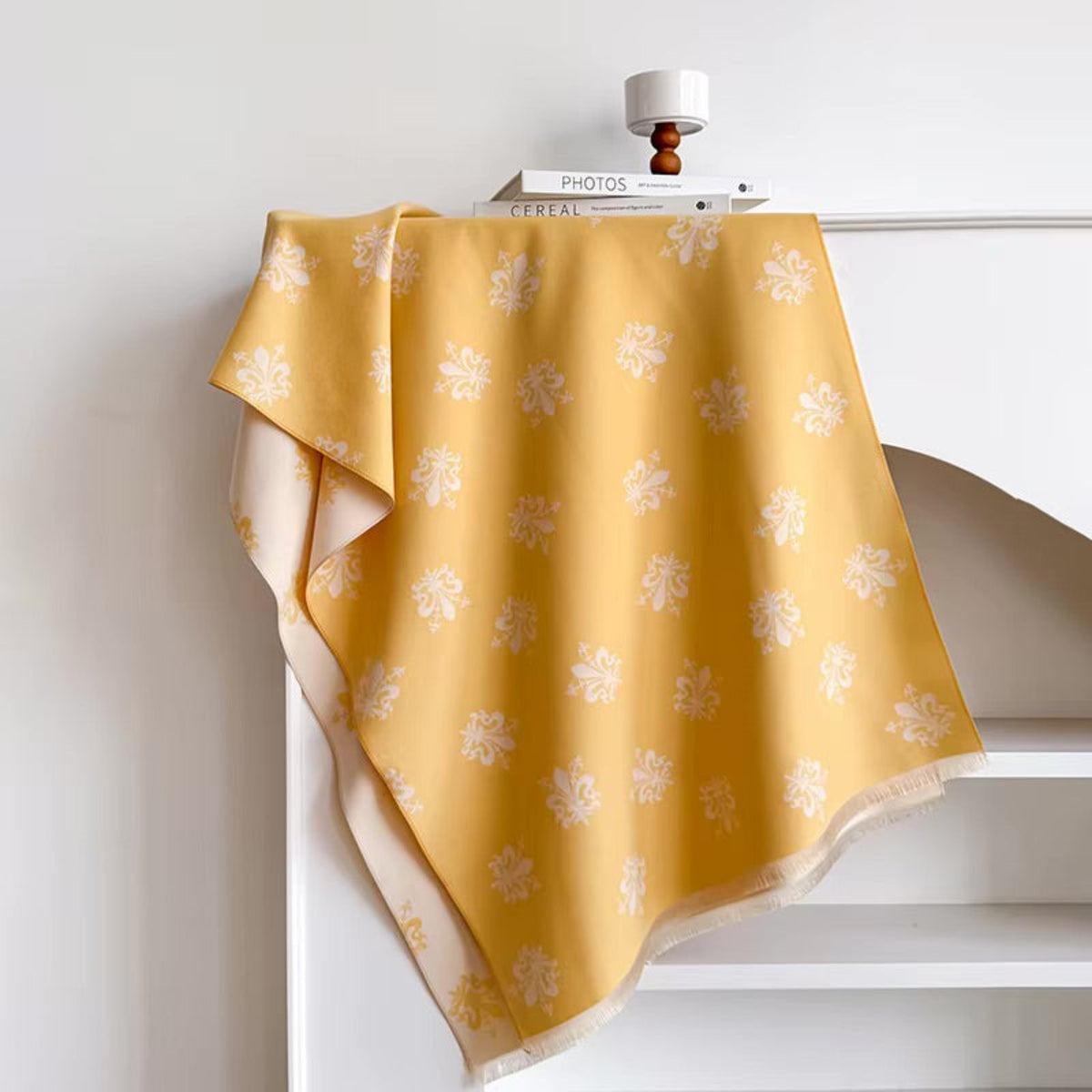 A mustard yellow fabric throw blanket with a white floral pattern draped over a white shelf, styled with a small white candle and books on top.