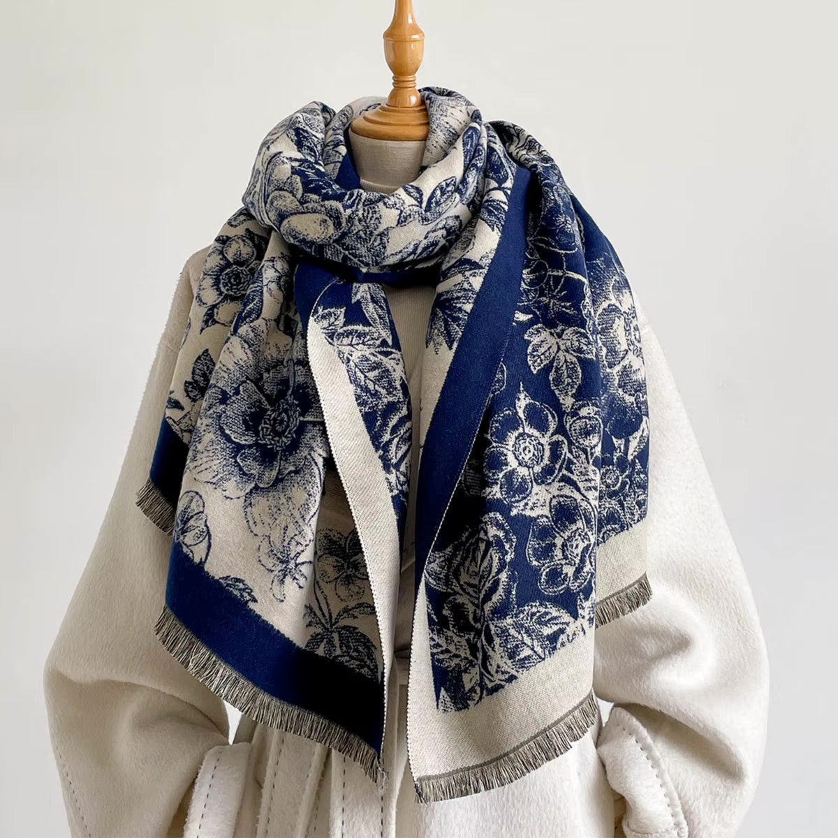 A navy blue and cream floral scarf with fringed edges, draped on a cream-colored coat displayed on a mannequin torso against a plain light background.