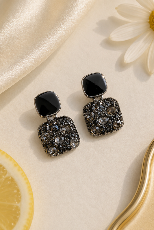 A pair of black drop earrings with square-shaped black stone tops and textured, rhinestone-encrusted square drops, styled on a cream satin surface with a daisy flower, a lemon slice, and a gold tray edge.