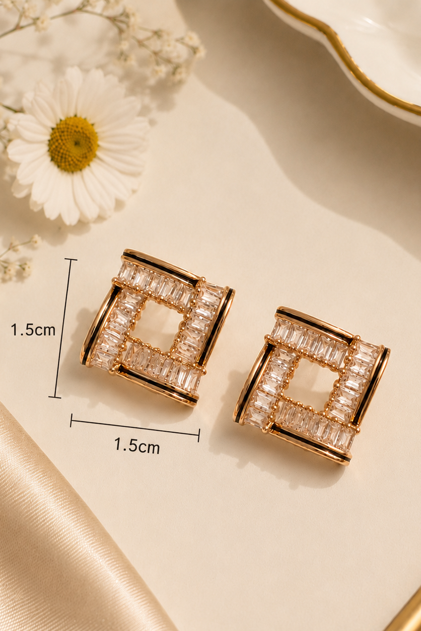A pair of geometric square stud earrings with clear rectangular stones in a rose gold-tone frame, shown on a beige surface with decorative dried flowers and a white dish. Measurement labels indicating 1.
