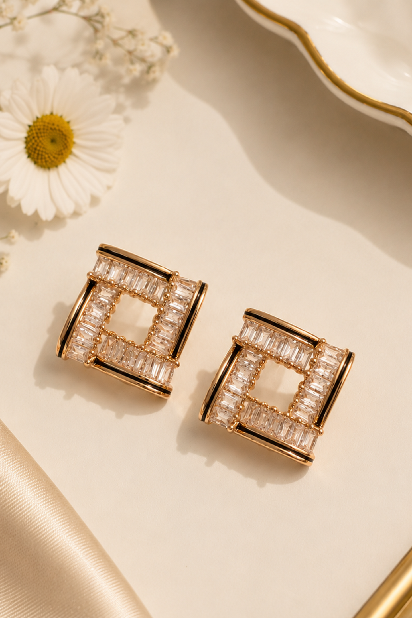 A pair of geometric square stud earrings with clear rectangular stones in a rose gold-tone setting, styled on a cream surface with a white daisy, dried flowers, satin fabric, and a white plate with a gold rim.