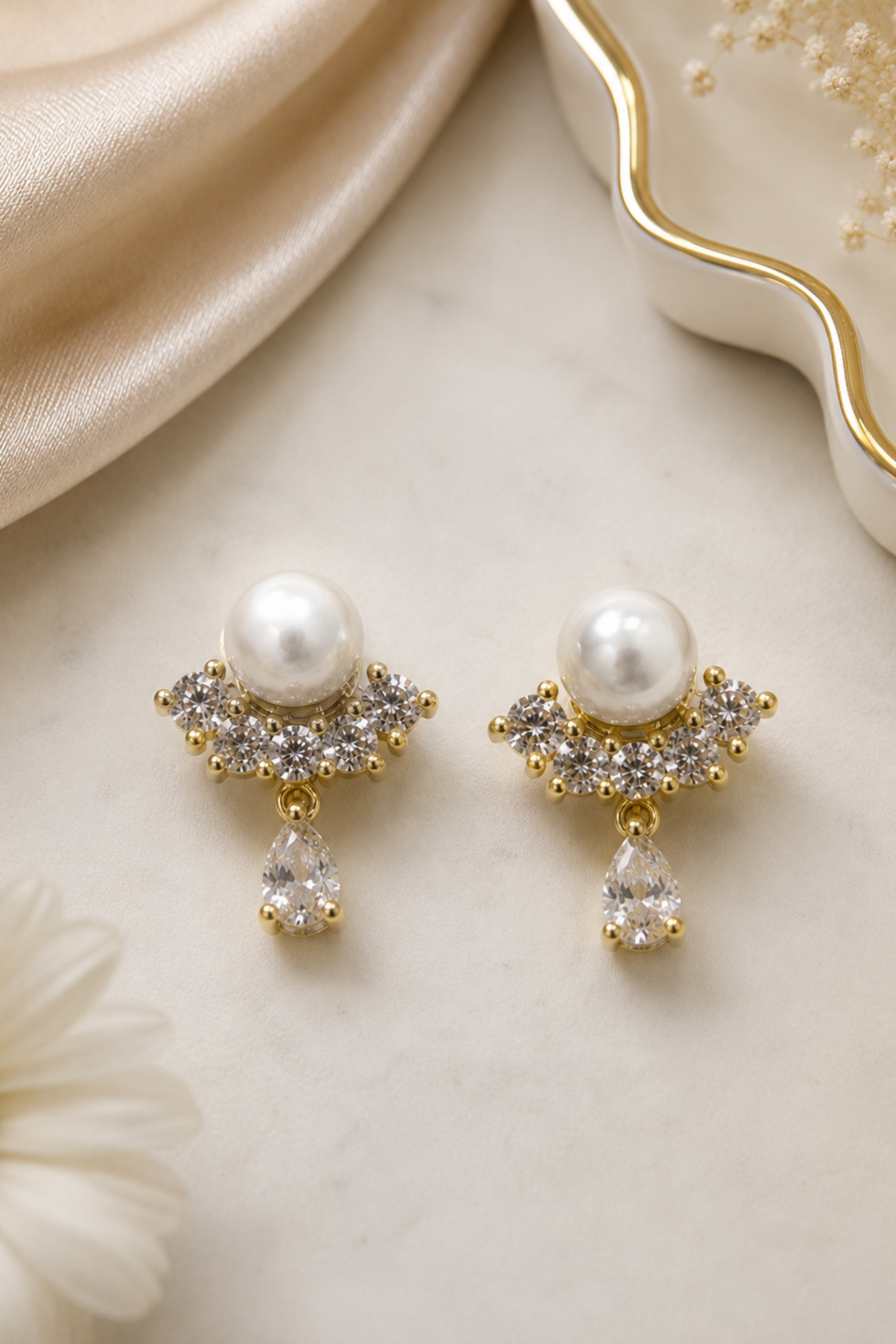 A pair of gold-plated drop earrings featuring a white pearl, a cluster of clear cubic zirconia stones, and a dangling teardrop-shaped clear stone, displayed on a light marble surface with gold and cream accents.