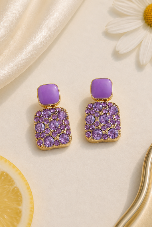 A pair of gold-tone drop earrings with a smooth lavender purple square top and a larger rectangular pavé-style lower section set with round lavender purple crystals, displayed on a cream surface with decorative fabric, a daisy, and a lemon slice.
