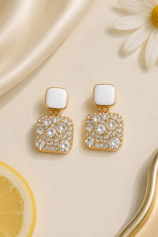 A pair of gold-tone drop earrings with a square white top and a larger square crystal-encrusted lower section, styled on a cream surface with satin fabric, a daisy flower, and a lemon slice.