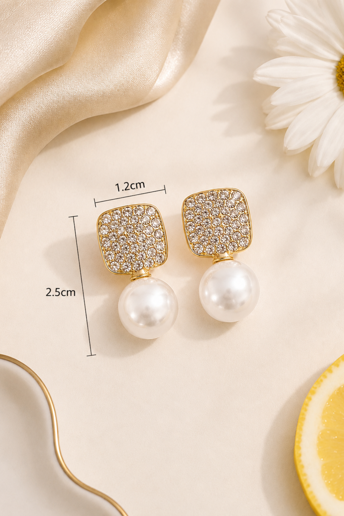 A pair of gold-tone drop earrings with square crystal-encrusted tops and round white pearl drops, shown on a light beige surface with decorative satin, a daisy flower, and a lemon slice. Black measurement lines and text indicate 1.