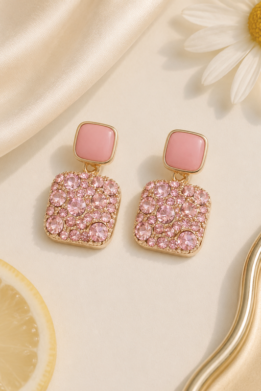 A pair of gold-tone drop earrings with square pink stone tops and larger pavé pink crystal square drops, styled on a cream satin surface with a daisy and lemon slice.
