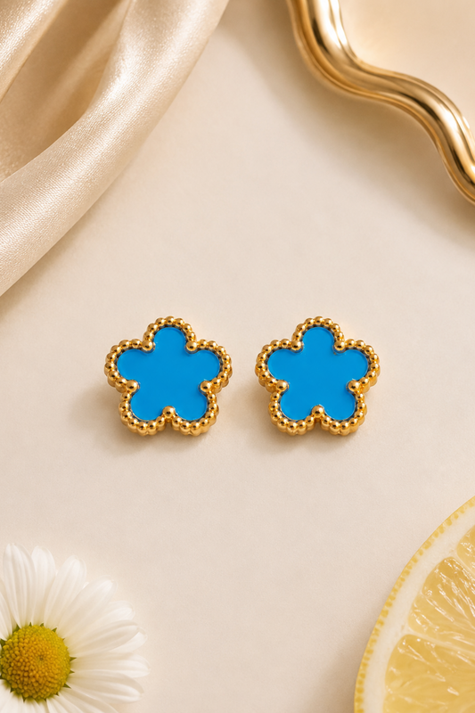 A pair of gold-tone flower-shaped stud earrings with bright blue enamel centers, styled on a light beige surface with a gold dish edge, draped satin, a daisy flower, and a lemon slice.