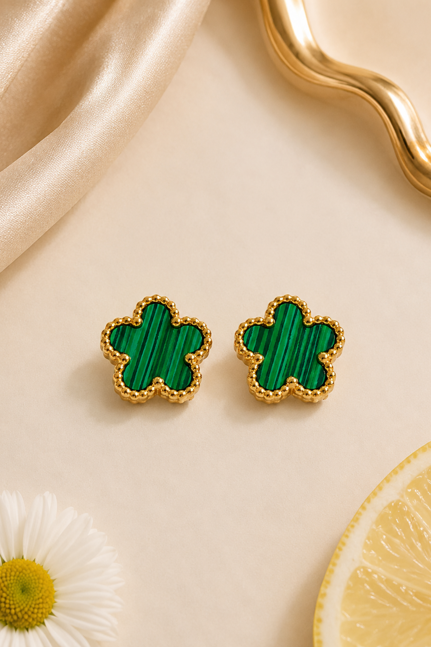 A pair of gold-tone flower-shaped stud earrings with green malachite inlay, styled on a beige surface with a satin fabric, a daisy flower, and a lemon slice.