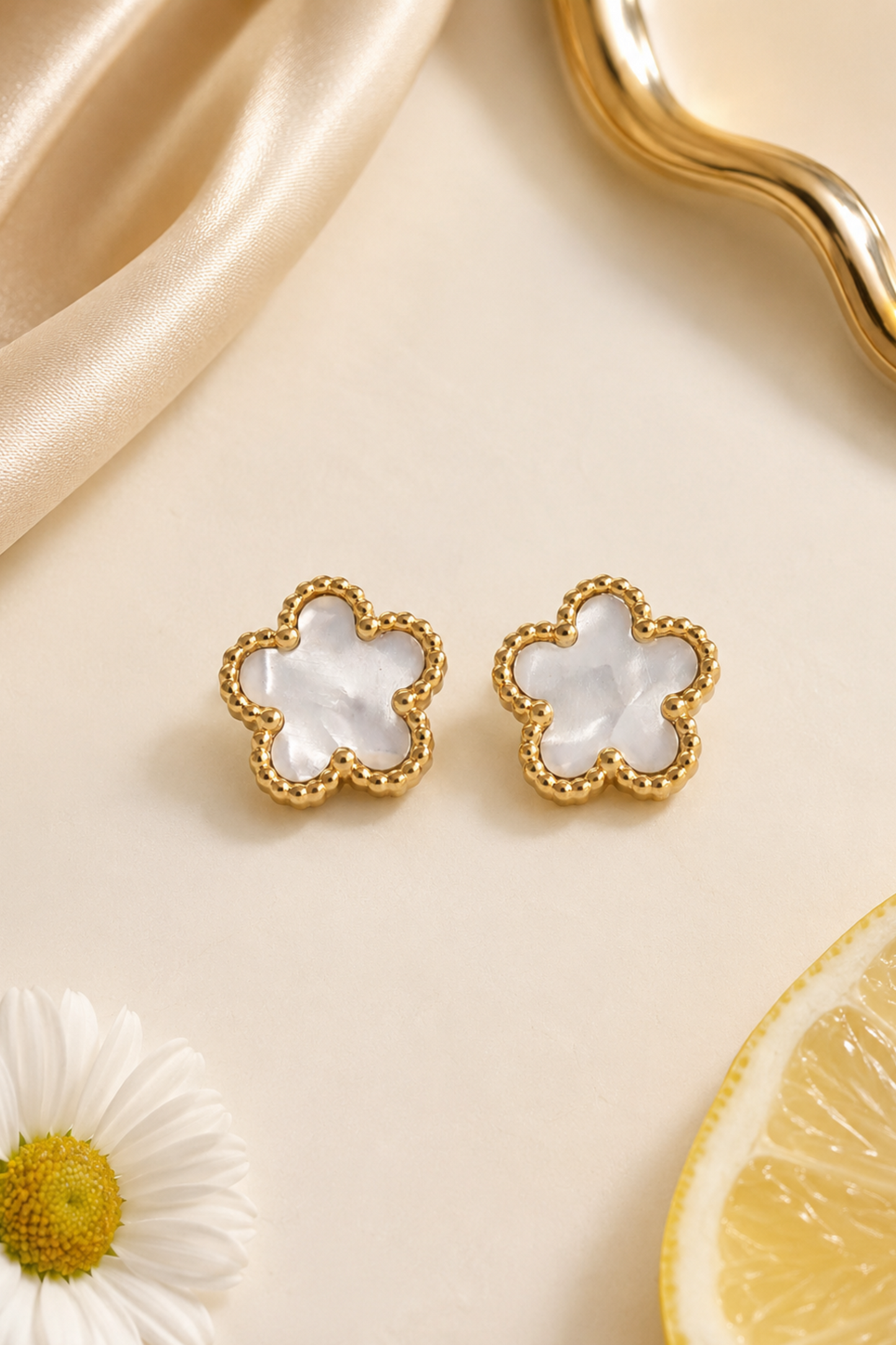 A pair of gold-tone flower-shaped stud earrings with white mother-of-pearl inlays, styled on a light beige surface with a gold wavy dish, satin fabric, a daisy flower, and a lemon slice.