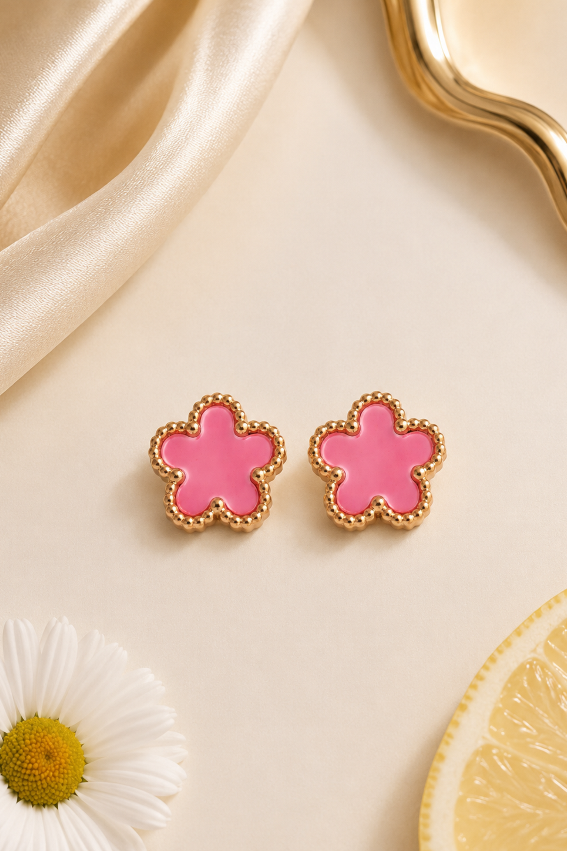 A pair of gold-tone flower stud earrings with a vibrant pink enamel center, styled on a cream background with a white daisy, a lemon slice, and draped champagne silk.