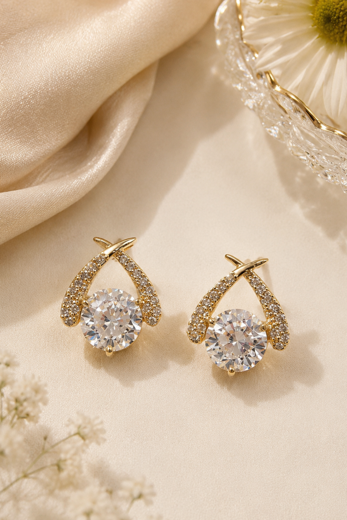 A pair of gold-toned drop earrings with a large clear round center stone and smaller clear stones along the curved upper arms, styled on a warm cream surface with satin fabric and a crystal dish.