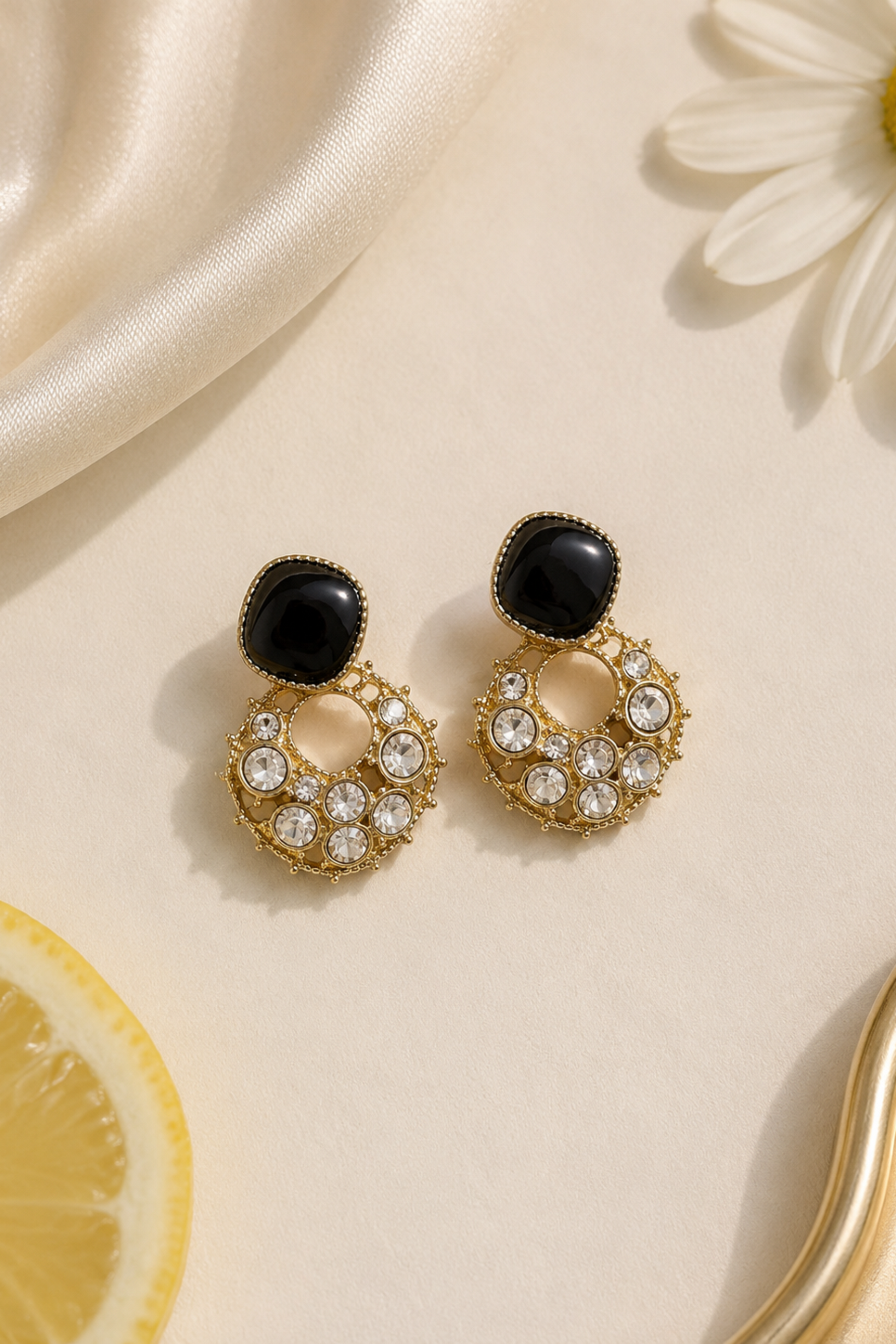 A pair of gold-toned drop earrings with black oval studs and clear rhinestone-covered crescent-shaped lower sections, styled on a cream surface with a daisy, satin fabric, lemon slice, and gold tray edge.