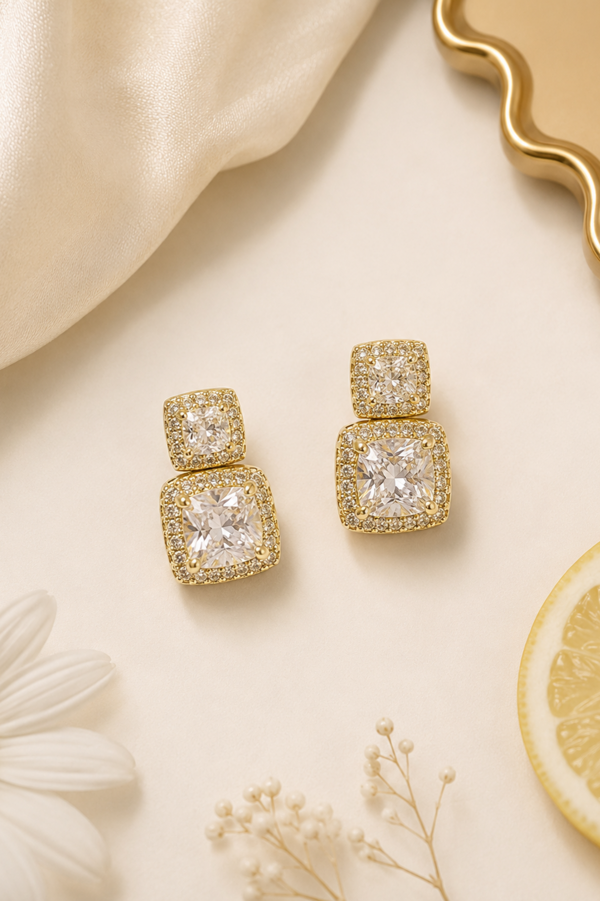 A pair of gold-toned drop earrings with square-cut clear stones and a halo of smaller clear stones, styled on a cream surface with a white daisy, dried flowers, a lemon slice, and draped satin fabric.