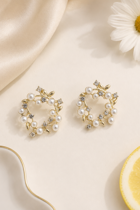 A pair of gold-toned floral stud earrings adorned with white faux pearls and small clear rhinestones, arranged on a cream surface with satin fabric, a daisy flower, and a lemon slice prop.