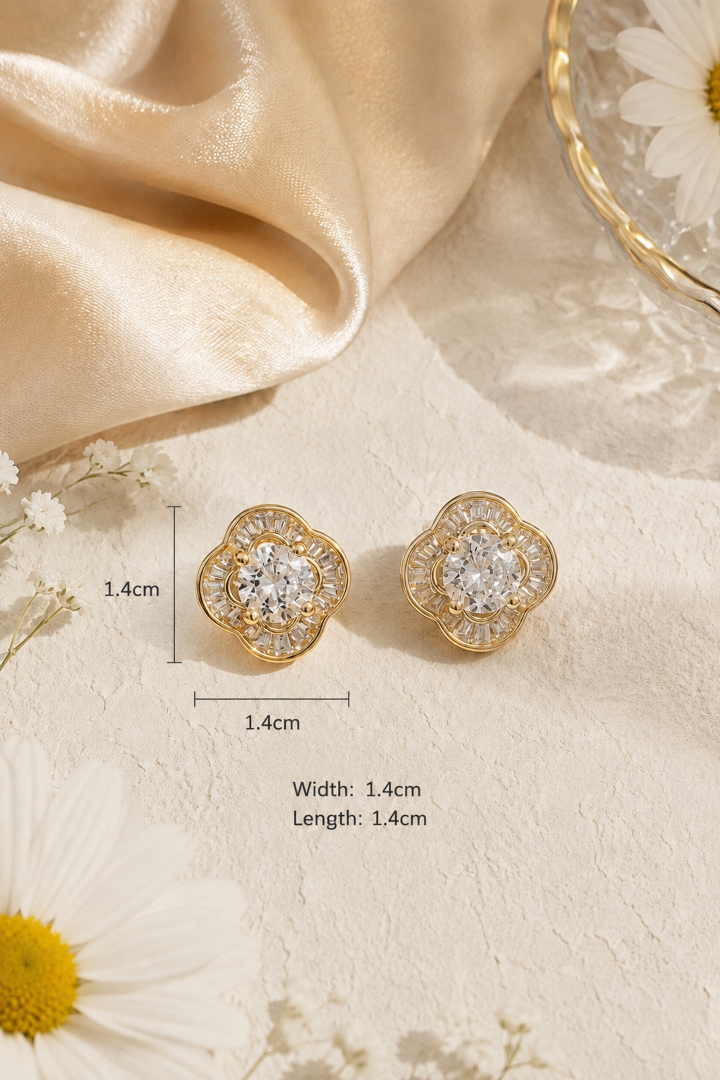A pair of gold-toned floral stud earrings with clear gemstones, shown on a textured cream fabric with draped satin and daisy props. Measurement text and arrows indicate the earrings are 1.